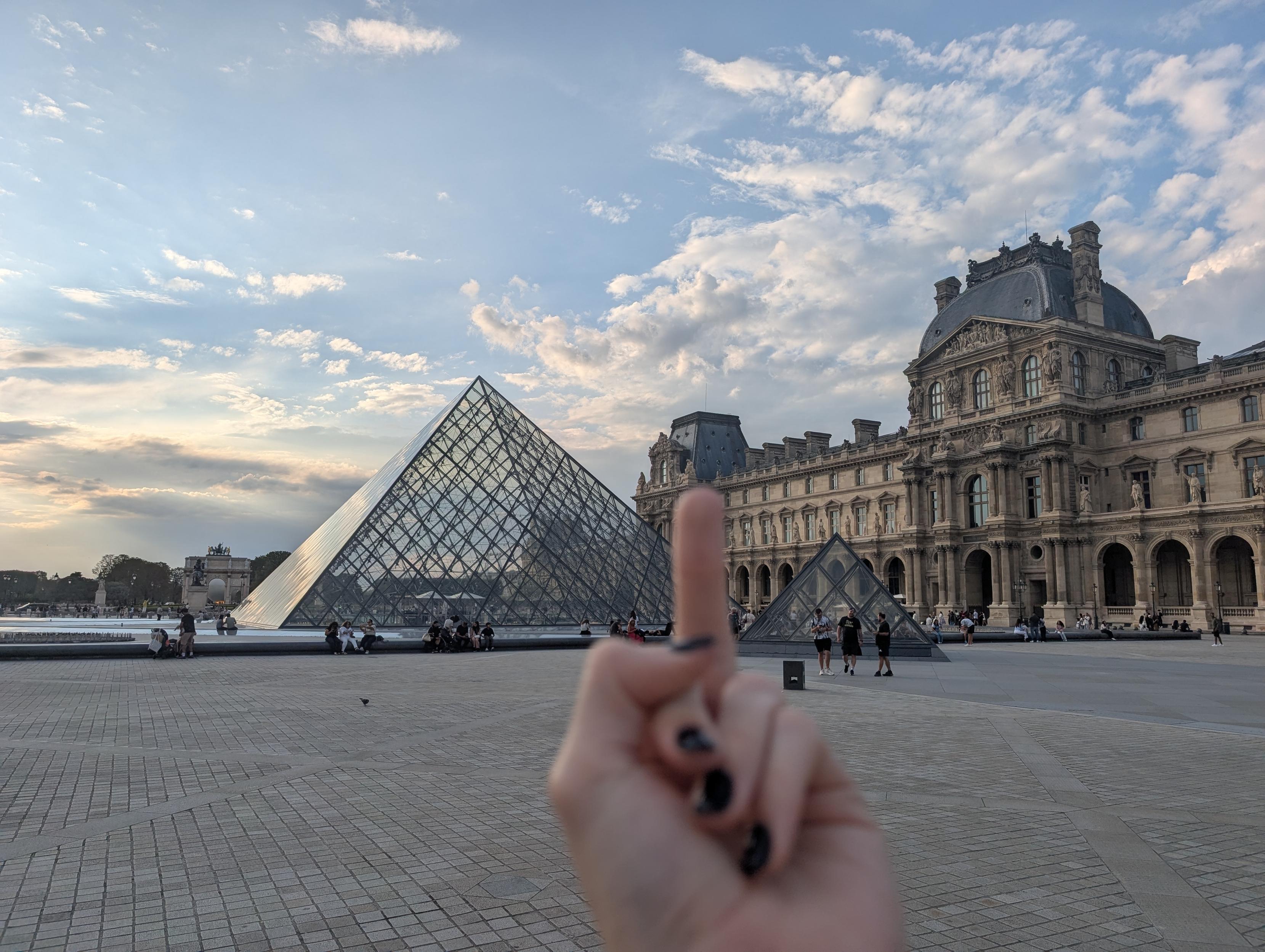 A photo of the pyramids outside the Louvre, with an out of focus middle finger pointed at them
